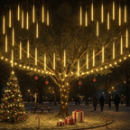 A festive outdoor Christmas scene with a large tree decorated with warm hanging light tubes and string lights, a lit Christmas tree at left, wrapped gift boxes at the base, and silhouetted people walking in the background at night.