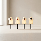 Four cute ghost-shaped solar garden lights with black stakes, glowing with a warm white light, arranged in a row on a light beige surface against a neutral beige wall.