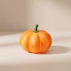 A small decorative orange pumpkin figurine with a green stem, standing on a light beige surface against a matching beige background with soft shadows.