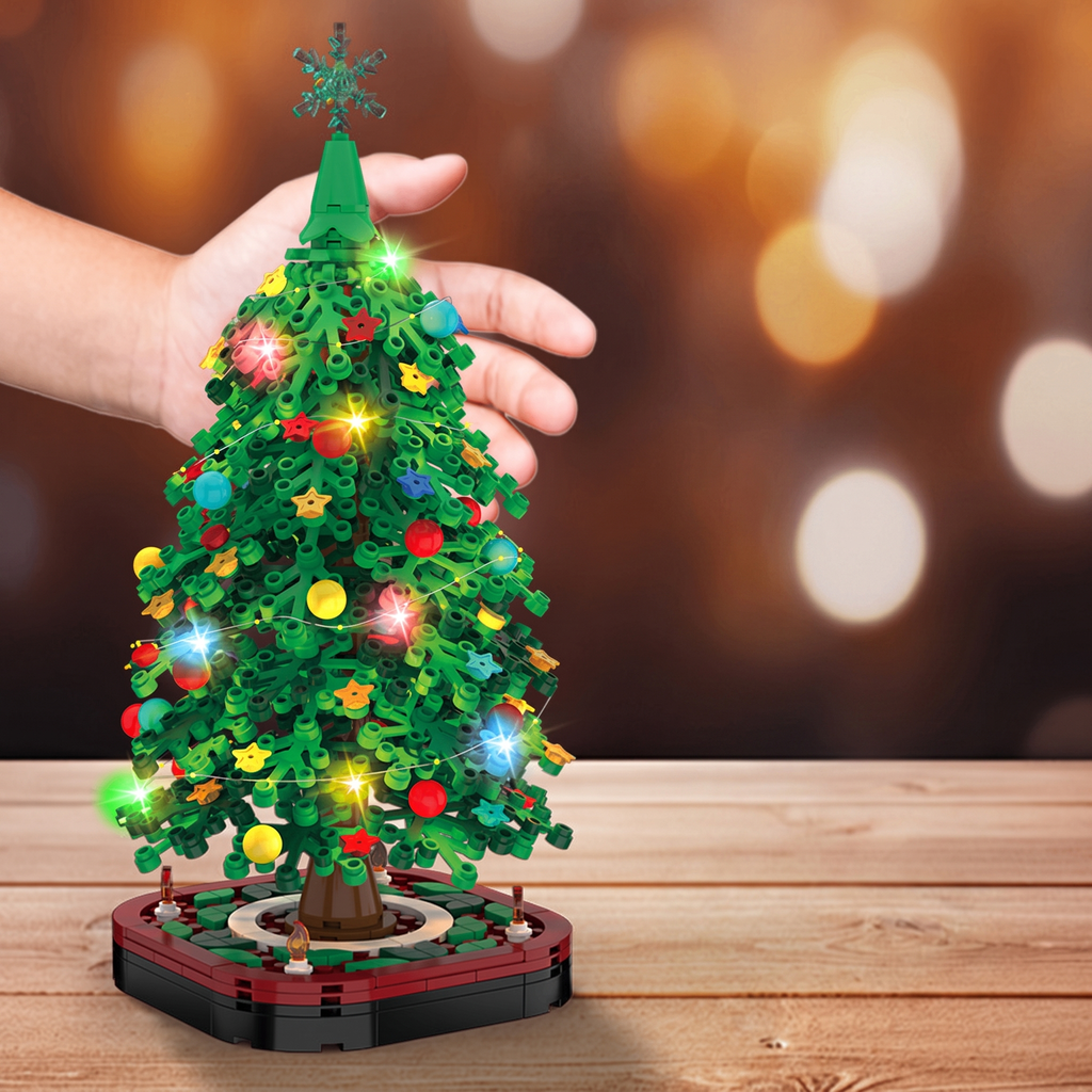A hand holds a small green LEGO-style Christmas tree with colorful ornaments and glowing lights on a wooden surface, against a warm blurred bokeh background.