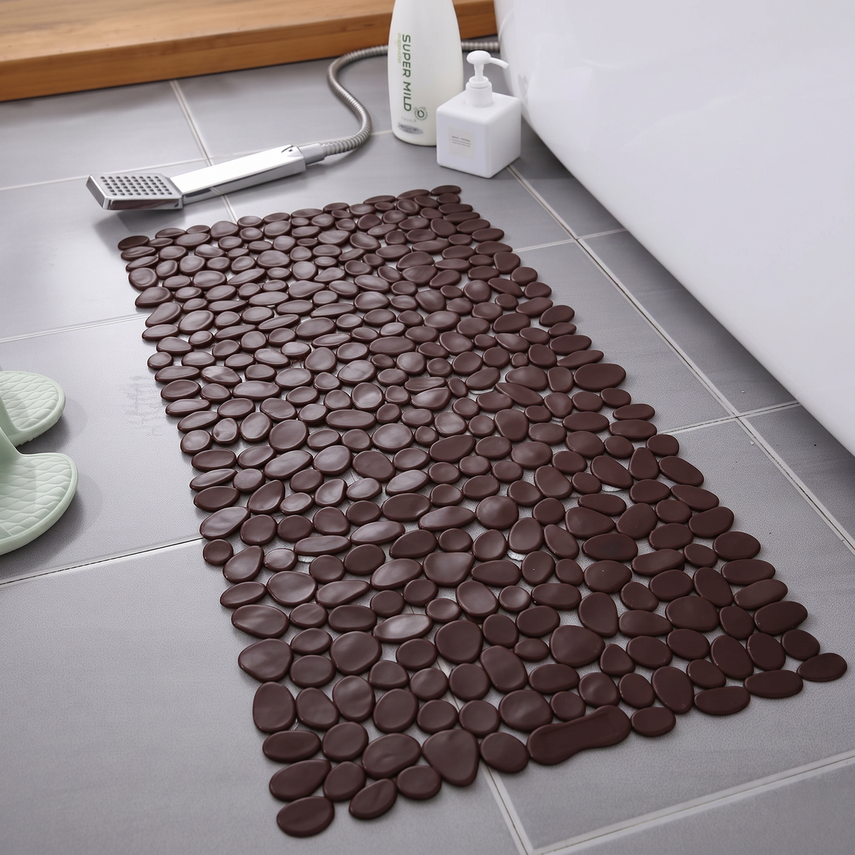 A dark brown pebble-patterned bath mat laid on a gray tiled bathroom floor next to a white bathtub, with a handheld shower head, soap dispenser, and a pair of light green slippers nearby.