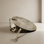 A surreal sculpture or Halloween decoration featuring a human skeleton reaching upward from a dark circular opening beneath a large, weathered oval stone slab, set in a minimalist beige room.