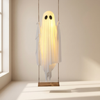 A ghost-shaped hanging swing lamp made from a glowing white sheet with black eyes and a wooden seat, suspended in a minimalist room by ropes.
