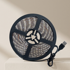 A coiled LED light strip with a black plastic reel and a USB connector, wound with small rectangular mosaic tiles in beige, white, and brown tones, set against a warm beige background.