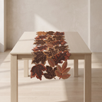 An autumn-themed table runner made of layered, realistic faux leaves in shades of brown, rust, and orange, displayed on a light wood dining table in a minimalist room.