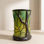 A sculptural ceramic cup or candle holder featuring a tree design with a green and yellow gradient background and dark brown branches and trunk, set against a warm beige background.