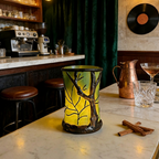 A decorative ceramic candle holder with a tree silhouette design, glowing with a warm yellow light, set on a marble counter in a cozy, dimly lit bar or cafe environment.