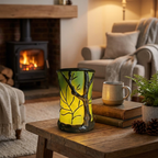 A glowing tree-bark patterned electric candle holder on a rustic wooden table in a cozy living room setting with a fireplace, armchair, and a ceramic mug.