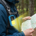 A close-up of a person outdoors in a forest wearing a blue hiking jacket and backpack, holding a map. A bright blue and black emergency light with a yellow LED panel is clipped to the chest strap.