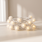 Coiled white dandelion string lights glowing with a warm yellow light, resting on a beige surface with soft window shadows.
