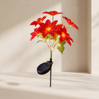 A solar-powered artificial poinsettia plant with glowing red flowers and green leaves, mounted on a black stake with an attached oval solar panel, set against a warm beige wall with soft shadows.
