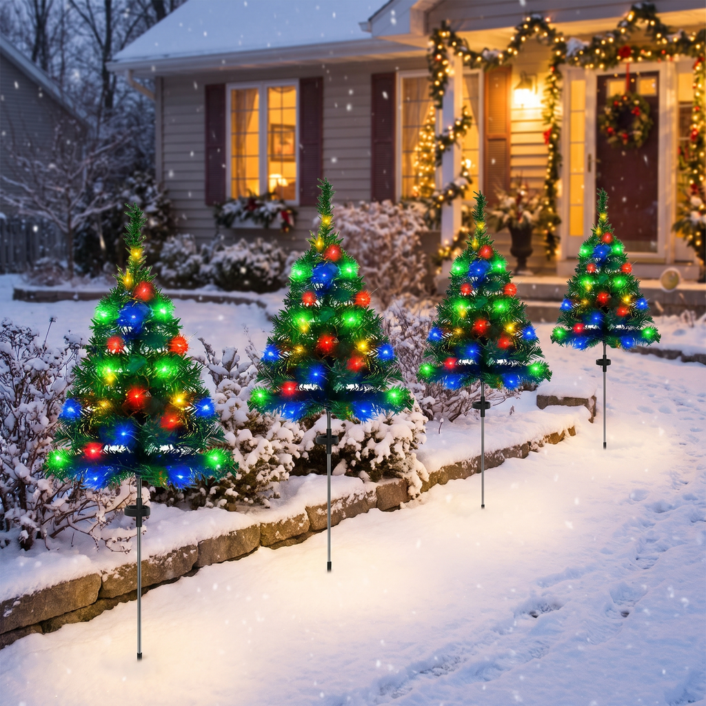 Four miniature LED Christmas tree yard lights in red, green, and blue, mounted on thin black stakes and arranged in a snowy front yard in front of a warmly lit, snow-covered house.