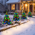 Four miniature LED Christmas tree yard lights in red, green, and blue, mounted on thin black stakes and arranged in a snowy front yard in front of a warmly lit, snow-covered house.
