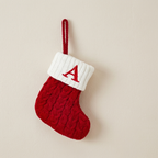 A small red and white knit Christmas stocking ornament with a textured cable-knit body, a white ribbed cuff, and a red embroidered letter 'A' on the cuff. It hangs from a red loop against a plain beige wall.
