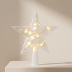 A clear acrylic star tree light with warm white LED fairy lights glowing inside, mounted on a white cylindrical base and displayed on a light beige surface against a matching neutral background.