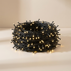 A coiled bundle of black string lights with warm white illuminated bulbs, photographed on a clean beige studio surface and background.