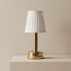 A table lamp with a white pleated fabric shade and a polished gold metal base, displayed on a matching cream-colored pedestal against a neutral beige background.