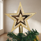 A large gold metallic star tree topper with layered star designs, displayed on a green Christmas tree branch against a neutral indoor wall.