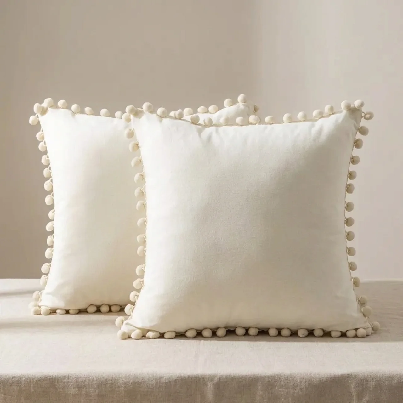 Two white pillows with pom-pom trim on a neutral background