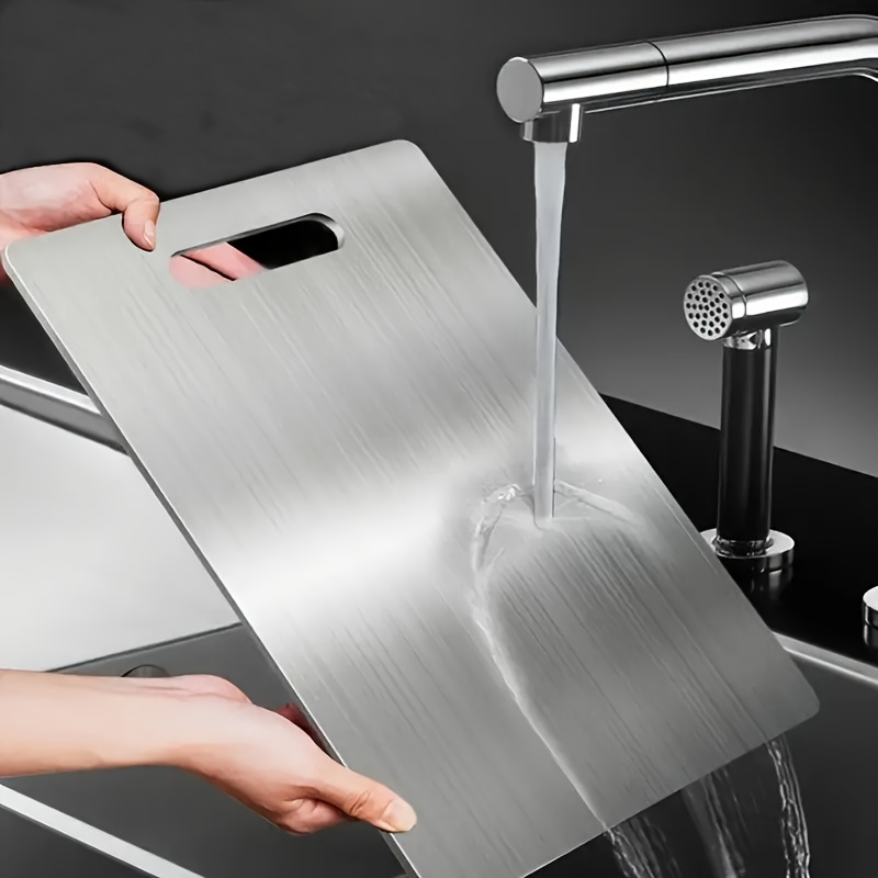 Person washing a metal cutting board under running water in a kitchen sink.