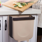 Beige wall-mounted trash bin next to a kitchen counter with a cutting board and vegetables.