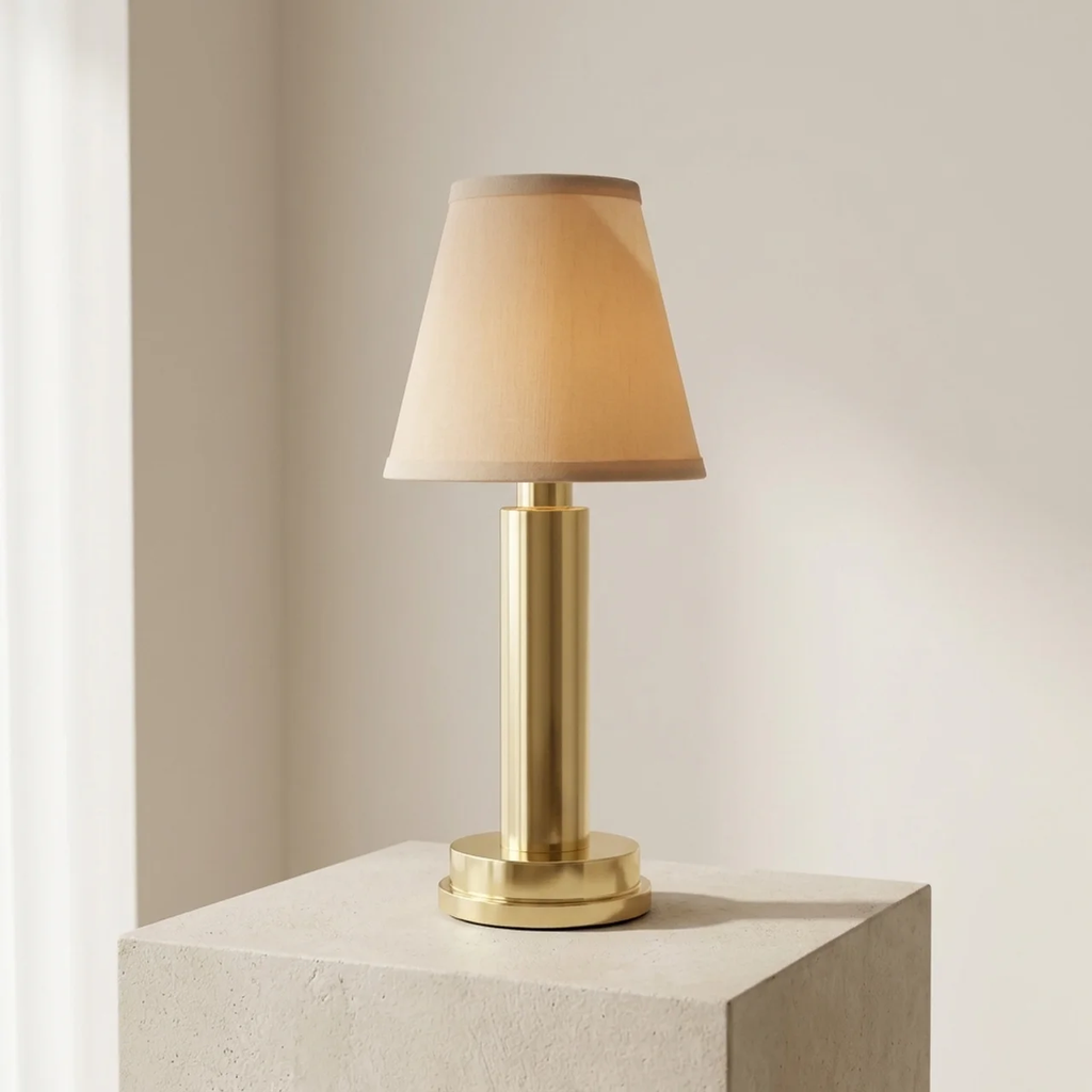 Gold table lamp with a beige shade on a white pedestal against a neutral wall.