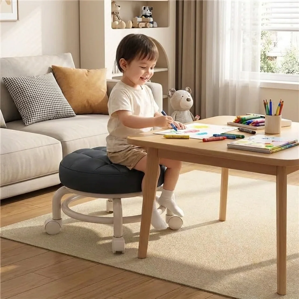 Child sitting on a black and white stool at a wooden table with art supplies in a living room.