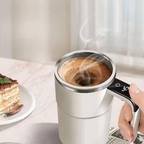 Person holding a white travel mug with steaming coffee next to a slice of cake on a light surface.