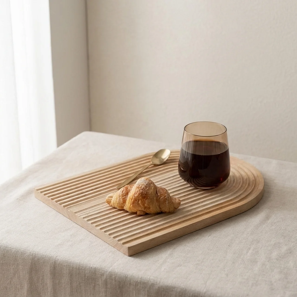 A light natural wood slatted serving board on a draped tablecloth, styled with a croissant, a spoon, and a glass of dark coffee in a softly lit interior setting.