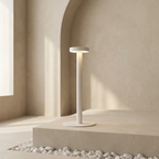 A minimalist white outdoor floor lamp with a circular shade and base, standing on a stone platform surrounded by white pebbles against a warm beige stucco wall.