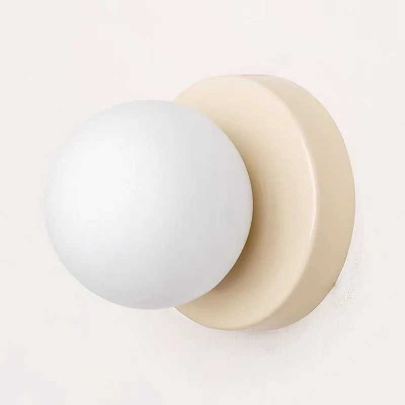 A minimalist wall-mounted light fixture with a matte white spherical globe and a warm cream-colored circular backplate, shown against a plain off-white wall.