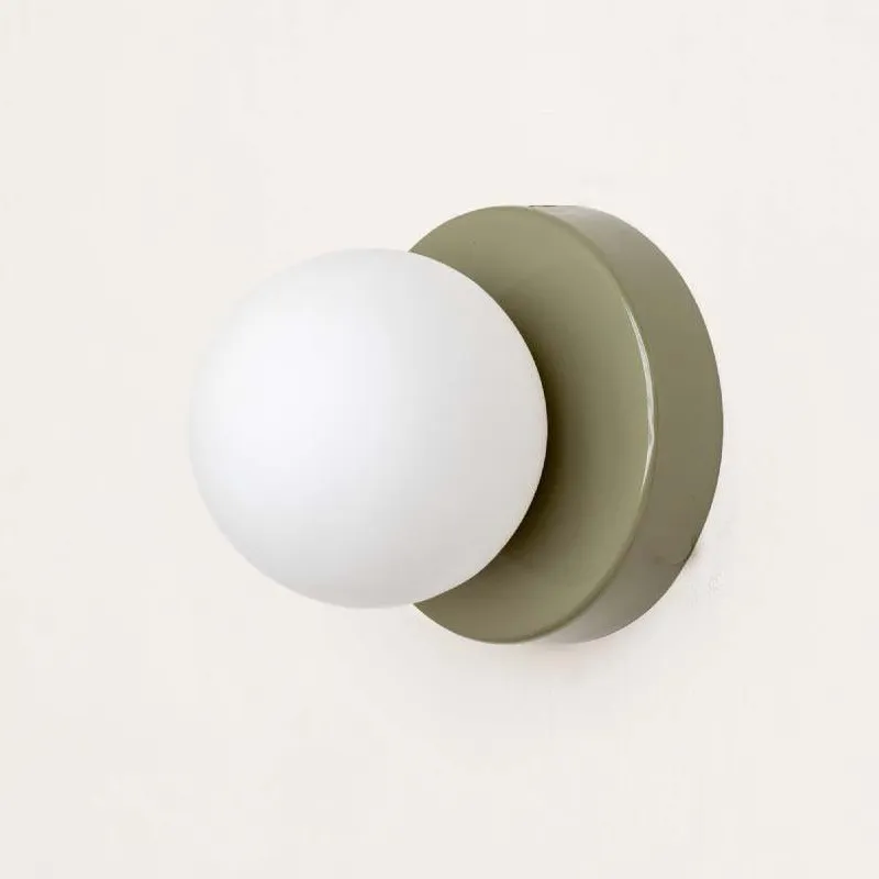 A minimalist wall-mounted light fixture featuring a matte white spherical globe shade and a muted sage green circular backplate, shown against a clean off-white background.