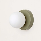 A minimalist wall-mounted light fixture featuring a matte white spherical globe shade and a muted sage green circular backplate, shown against a clean off-white background.