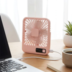 A small pink portable desk fan with a USB charging port, shown plugged in on a wooden table next to a laptop, a cup of coffee, and a potted plant.
