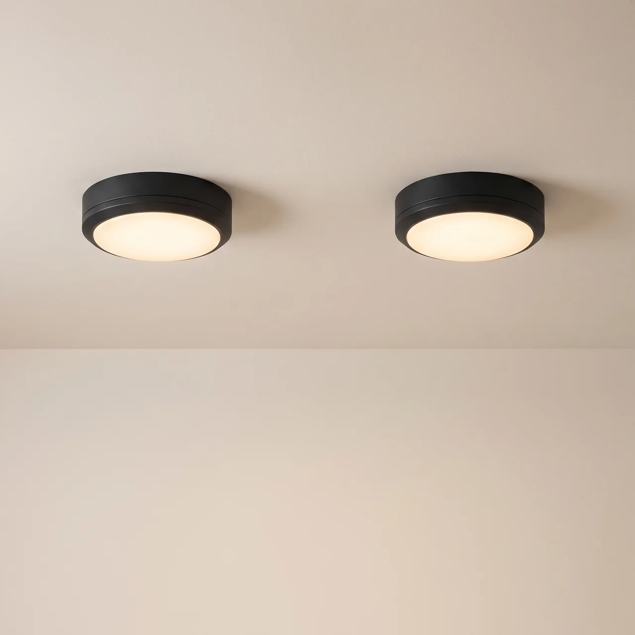 Two modern flush-mount ceiling lights with matte black circular frames and warm white glowing diffusers, installed side-by-side on a beige ceiling.