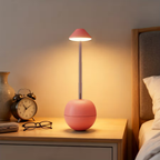 A pink spherical table lamp with a slim metallic stem and a pink conical shade, glowing warm white on a wooden bedside table next to a clock, glasses, and a bed.