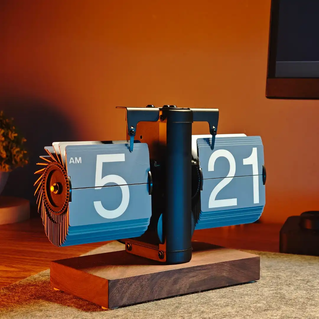 A retro-style mechanical flip clock with blue numbers on a wooden base, photographed in a warm indoor setting with an orange wall and carpet.