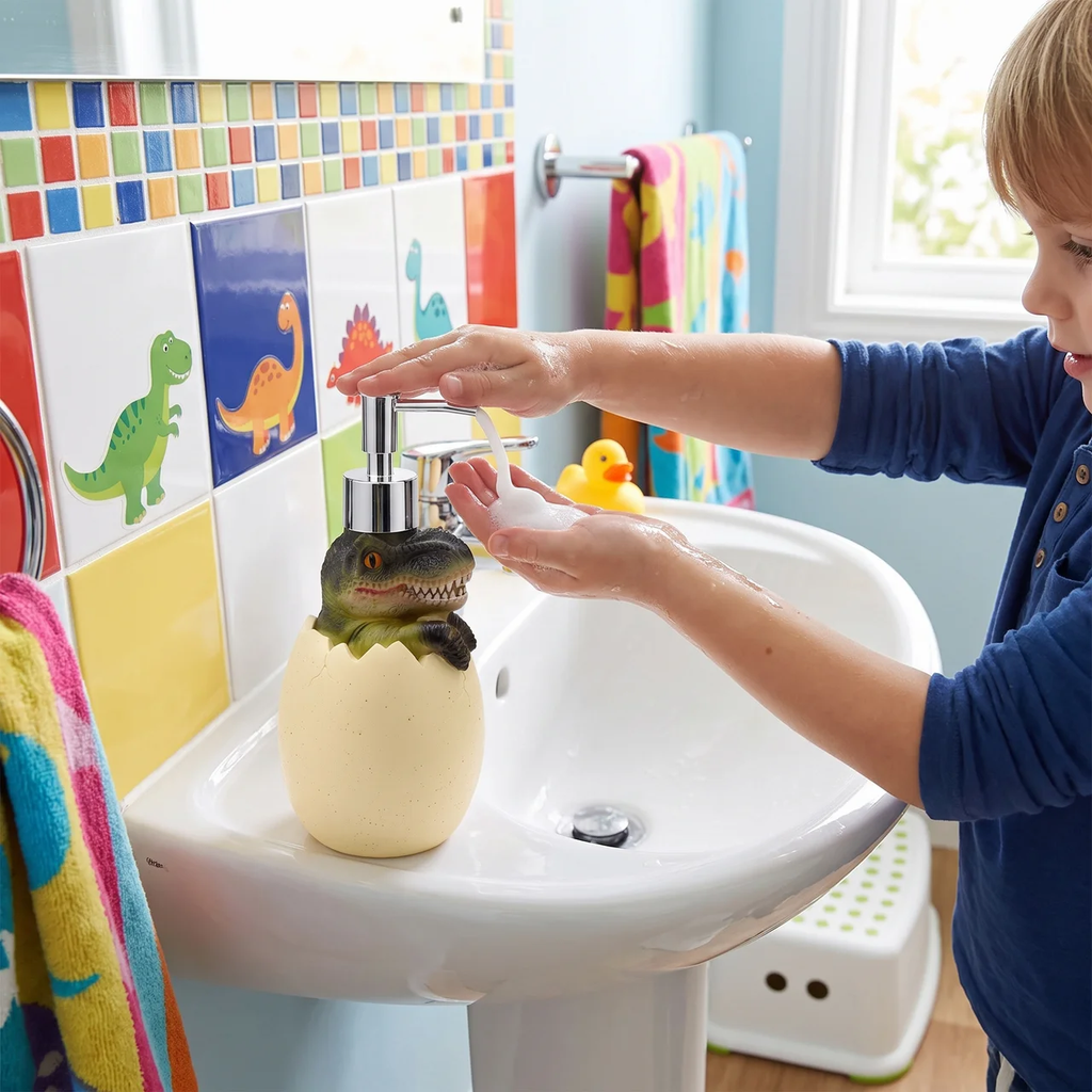 A child washing their hands at a white pedestal sink, using a dinosaur-themed soap dispenser that hatches an egg. The sink is set against a colorful tiled wall with dinosaur illustrations.