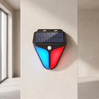 A triangular solar-powered outdoor light fixture mounted on a beige wall, featuring a black top housing a solar panel, a central white sensor, and two glowing light panels in blue and red below.