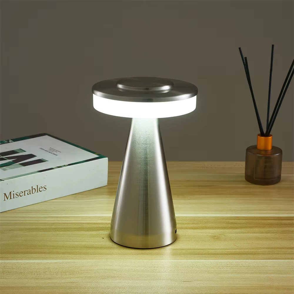 A modern table lamp with a brushed metal cone-shaped base and a circular light-up top, shown lit on a wooden tabletop with a book and reed diffuser in the background.