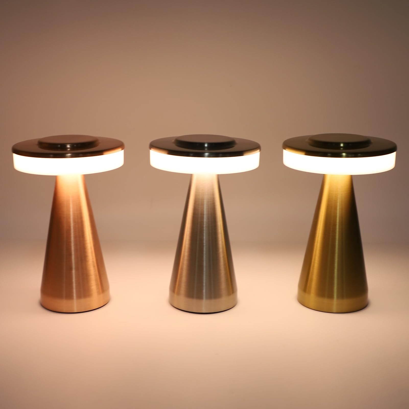 Three modern table lamps with brushed metal cone-shaped bases and glowing white circular light bands, shown in copper, silver, and gold finishes against a warm beige background.