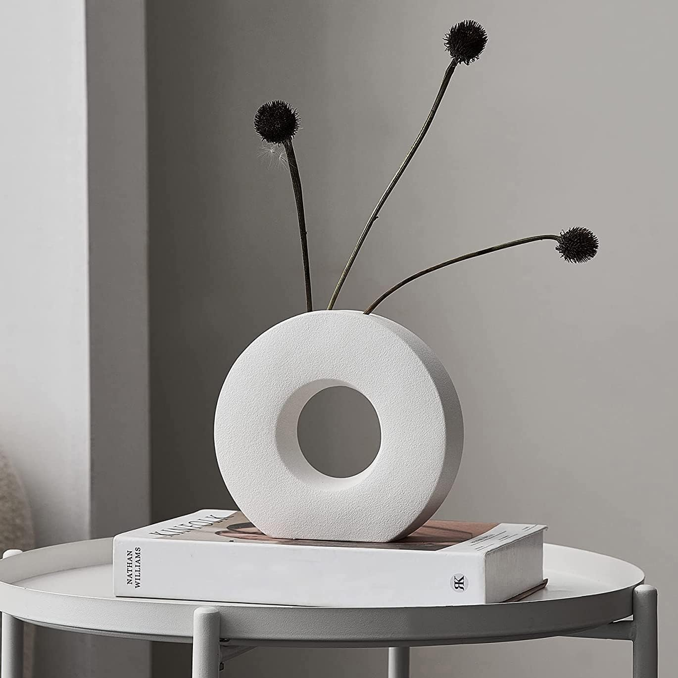A white donut-shaped ceramic vase with a circular center cutout, holding three long black stems with round dark flower heads, displayed on a white round table with a book underneath against a soft gray wall.