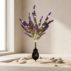 Purple artificial flower stems with embedded warm white fairy lights in a black lantern-shaped vase, displayed on sand with small stones against a beige wall near a window.