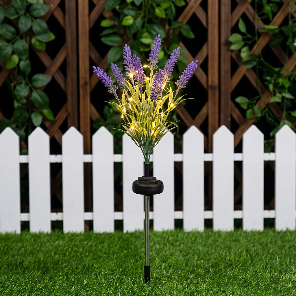 Solar-powered artificial lavender garden stake with glowing warm white lights, set in green grass in front of a white picket fence and wooden trellis.