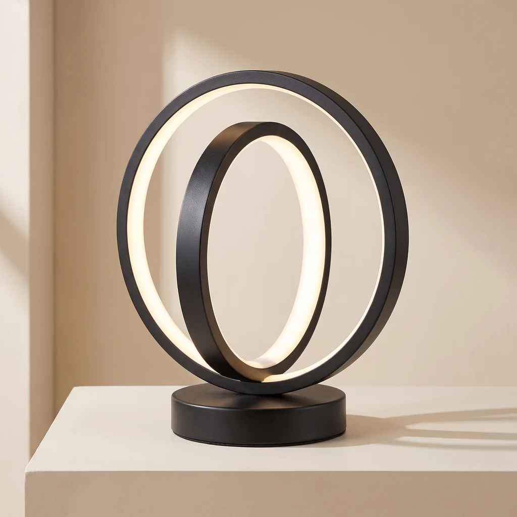 A modern black metal table lamp with two interlocking circular rings, one illuminated with a warm white glow, set on a matching round base.