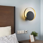 A modern circular wall sconce with a gold outer ring and a dark gray center, mounted on a light blue-gray wall above a bed in a bedroom setting.
