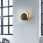 A modern wall-mounted light fixture with a gold-toned circular frame and a dark gray central disc, shown lit on a white wall next to black-framed windows.