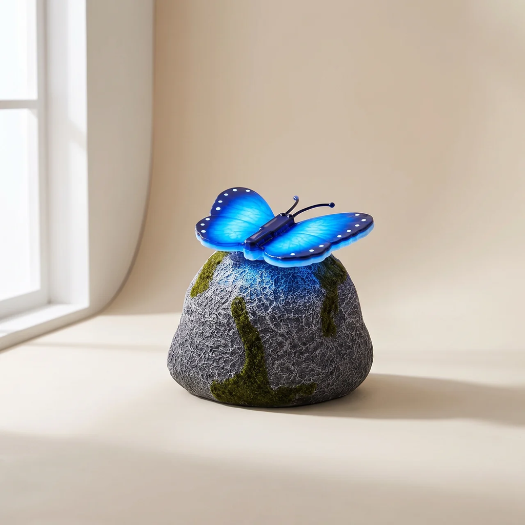 A decorative garden light shaped like a mossy rock with a glowing blue butterfly perched on top, photographed on a warm beige surface near a window.