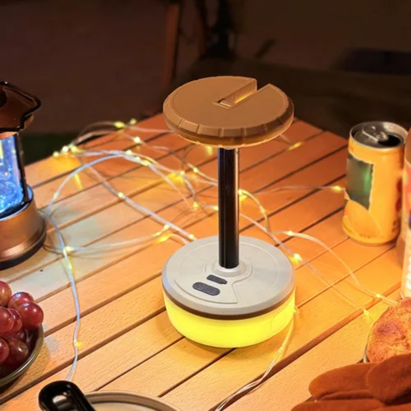 A portable camping lantern with a circular top lid and a glowing yellow base, placed on a wooden table outdoors at night with string lights, a beverage can, and food nearby.