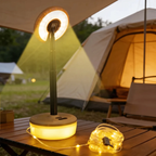 A camping table lamp with a circular light head and glowing cylindrical base sits on a wooden table outdoors near a tent. A coiled string of warm white fairy lights is placed beside it, with the lamp casting a yellow-green light beam into the evening campsite setting.